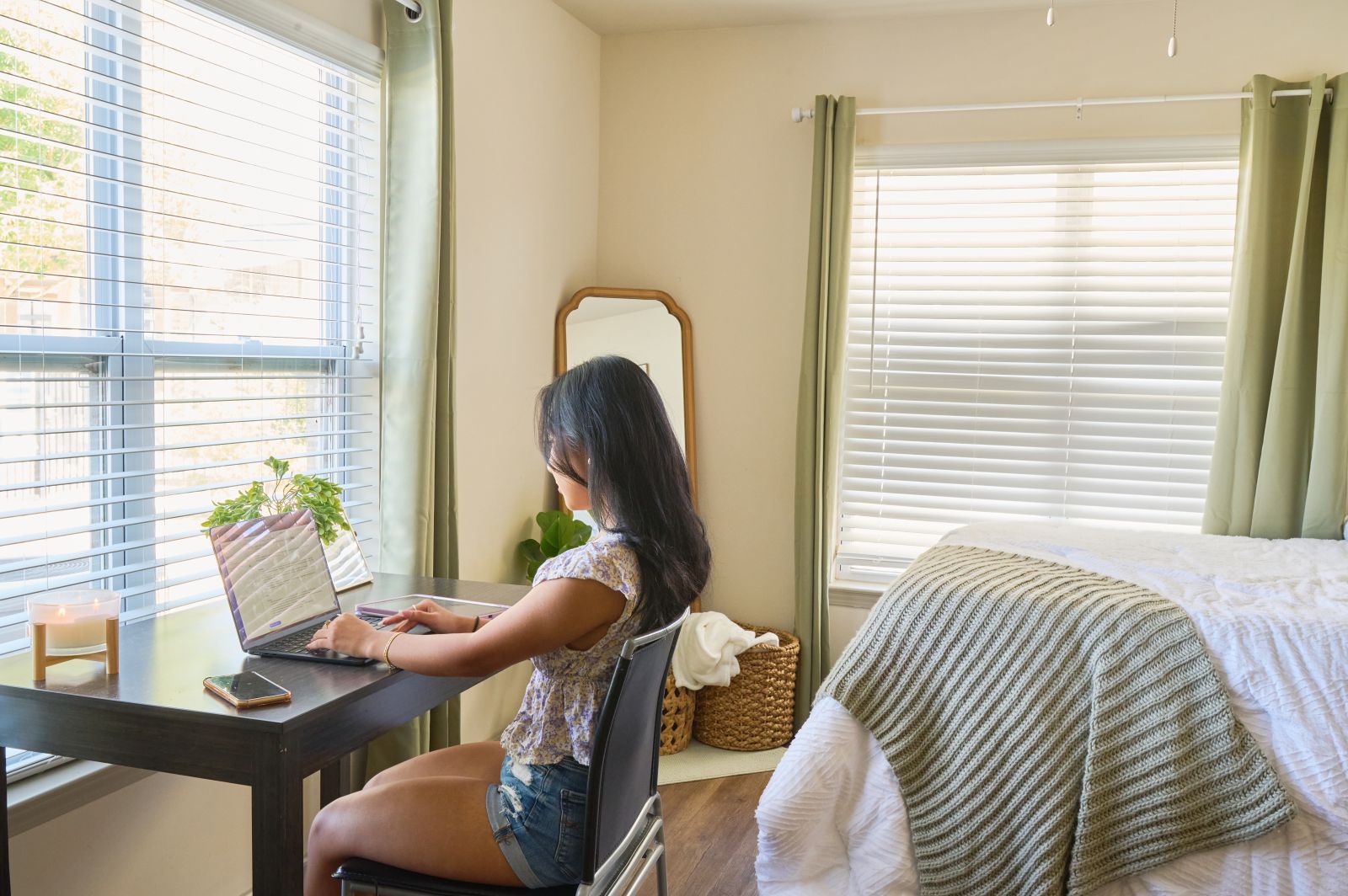 A woman seated at a desk with a bed visible in the background engaged in work or contemplation